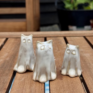 May include: Three small ceramic ghost figurines in varying heights, each featuring a smooth, off-white glazed finish with subtle tan shading around the edges, pointed ears, and two simple circular indentations for eyes on their front faces.