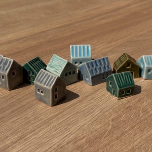 May include: A collection of ten miniature ceramic houses in various shades of blue, green, and grey. Each small figurine features a gabled roof with vertical ridges, square windows, and a simple door, arranged in a cluster on a flat surface.