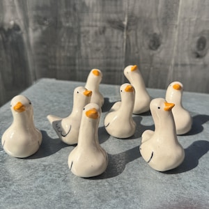 May include: A collection of seven small, off-white ceramic duck figurines. Each duck features a smooth, rounded body, a bright orange beak, and a single black dot for an eye. The figures are arranged in a group, showcasing their simple, hand-crafted form.