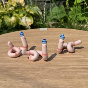 May include: Three light pink ceramic worm figurines, each featuring a small blue and white striped party hat on its head. The worms have simple black dot eyes and a curved mouth, with a single pink band around their bodies and coiled tails.