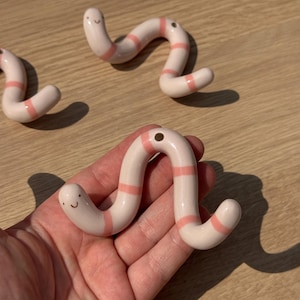 May include: A light pink ceramic worm figurine with horizontal coral-coloured stripes and a small, simple hand-painted face. The worm has a curved, segmented body shape and a small circular hole near one end, featuring a smooth, glossy finish.