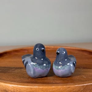 以下が含まれることがあります： 2つのセラミックハトの置物。鳥は主にグレーで、体に紫と緑のアクセントがあります。小さな黒いクチバシと目があります。置物は丸い木製のトレイに飾られています。