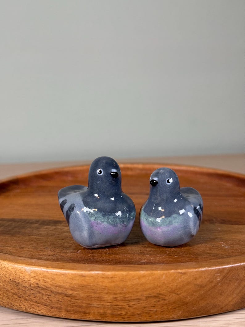 以下が含まれることがあります： 2つのセラミックハトの置物。鳥は主にグレーで、体に紫と緑のアクセントがあります。小さな黒いクチバシと目があります。置物は丸い木製のトレイに飾られています。