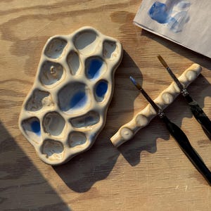 May include: A ceramic paint palette with irregular, organic-shaped wells featuring white and blue pigment residue, accompanied by a matching ceramic brush rest holding two paintbrushes with black handles and metal ferrules.
