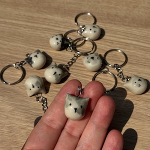 May include: A collection of small, handmade ceramic cat head charms attached to silver-toned metal keychains. Each charm features a smooth, off-white glazed finish with hand-painted grey patches, black eyes, and a small black nose detail.
