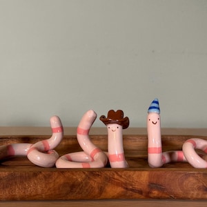 May include: Three light pink ceramic worms with darker pink horizontal stripes and simple painted faces. One worm wears a brown cowboy hat, and another wears a small blue and white striped party hat. The worms are arranged in a wooden tray.