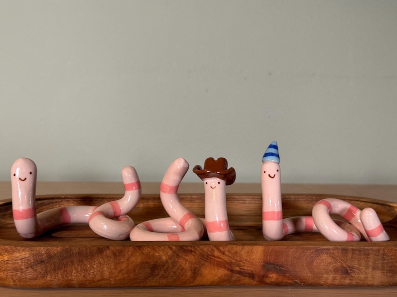 May include: Three light pink ceramic worms with darker pink horizontal stripes and simple painted faces. One worm wears a brown cowboy hat, and another wears a small blue and white striped party hat. The worms are arranged in a wooden tray.