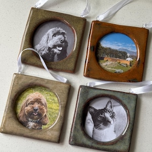 May include: Four square ceramic frames with glossy glazes in shades of olive, brown, and sage green. Each frame features a circular centre cutout displaying a photograph and includes a white ribbon loop at the top for hanging purposes.