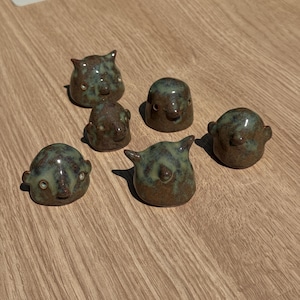 May include: A collection of six small, hand-sculpted ceramic figurines with rounded shapes. Each piece features a mottled glaze in shades of earthy brown and sage green, with simple, embossed facial features including small eyes and noses.