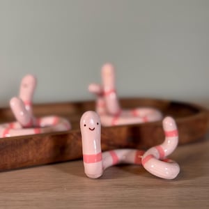 May include: Three small, glossy ceramic worm figurines in a light pink color with horizontal coral-pink stripes. Each worm has a simple, hand-painted face with two small black dots for eyes and a curved line for a mouth, positioned in a coiled pose.