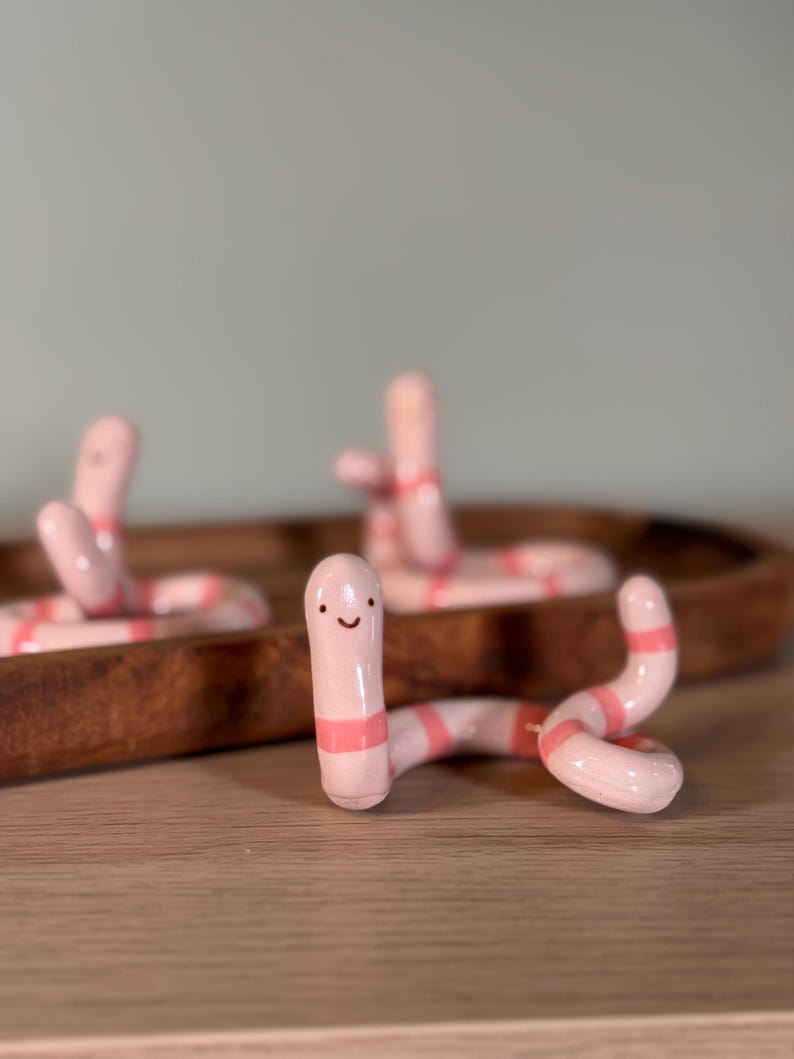 May include: Three small, glossy ceramic worm figurines in a light pink color with horizontal coral-pink stripes. Each worm has a simple, hand-painted face with two small black dots for eyes and a curved line for a mouth, positioned in a coiled pose.