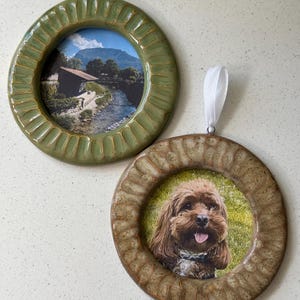 May include: Two circular ceramic ornaments with white ribbon loops for hanging. One frame is sage green and contains a landscape photo of a river and mountain. The other frame is tan with a textured border and features a photo of a brown dog.