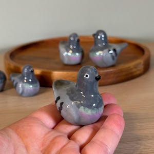 以下が含まれることがあります： 陶器製のハトの置物のコレクション。鳥は主にグレーで、紫と緑の虹色のアクセントがあります。1つの置物が手に持たれ、他の置物は木製のトレイに配置されています。ハトは黒い嘴をしています。