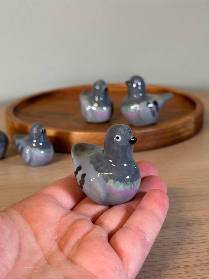 以下が含まれることがあります： 陶器製のハトの置物のコレクション。鳥は主にグレーで、紫と緑の虹色のアクセントがあります。1つの置物が手に持たれ、他の置物は木製のトレイに配置されています。ハトは黒い嘴をしています。