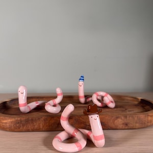 May include: Four small, light pink ceramic worms with darker pink horizontal stripes and simple painted faces. One worm wears a brown cowboy hat, another wears a small blue and white striped party hat, and two are plain.