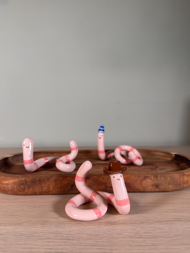 May include: Four small, light pink ceramic worms with darker pink horizontal stripes and simple painted faces. One worm wears a brown cowboy hat, another wears a small blue and white striped party hat, and two are plain.