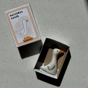 May include: A small white ceramic goose figurine with an orange beak and black eye details, resting inside a cardboard matchbox. The box lid features a graphic illustration of a goose with the text MATCHBOX GOOSE and the brand name SAMRAMICS.