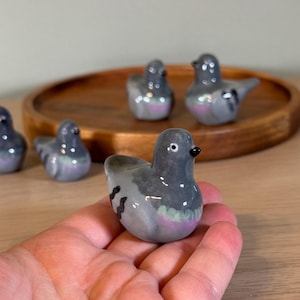 以下が含まれることがあります： 小さなセラミック製のハトの置物のコレクション。鳥は主に灰色で、胸と羽に虹色の紫と緑のアクセントがあります。各置物には黒いくちばしと小さくて丸い目があります。木製のトレイに配置されているものもあります。