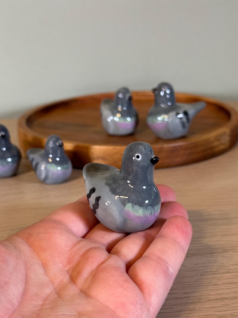 以下が含まれることがあります： 小さなセラミック製のハトの置物のコレクション。鳥は主に灰色で、胸と羽に虹色の紫と緑のアクセントがあります。各置物には黒いくちばしと小さくて丸い目があります。木製のトレイに配置されているものもあります。