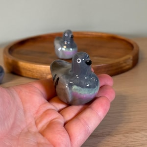 以下が含まれることがあります： グレーの体、黒い嘴、ピンクと緑の虹色のアクセントが施されたセラミック製のハトの置物。鳥は手で持たれており、木製のトレイには他の2つの同様の置物が見えます。ハトの置物は装飾品です。