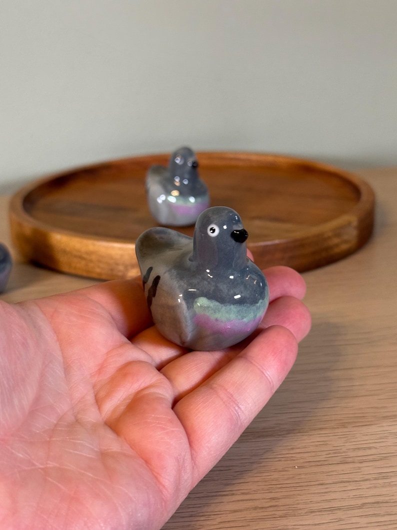 以下が含まれることがあります： グレーの体、黒い嘴、ピンクと緑の虹色のアクセントが施されたセラミック製のハトの置物。鳥は手で持たれており、木製のトレイには他の2つの同様の置物が見えます。ハトの置物は装飾品です。
