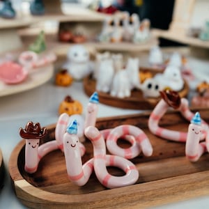 May include: A collection of small, hand-sculpted ceramic worms in light pink with darker pink stripes. Each worm features a simple smiling face and wears a tiny accessory, such as a brown cowboy hat or a blue and white patterned party hat.