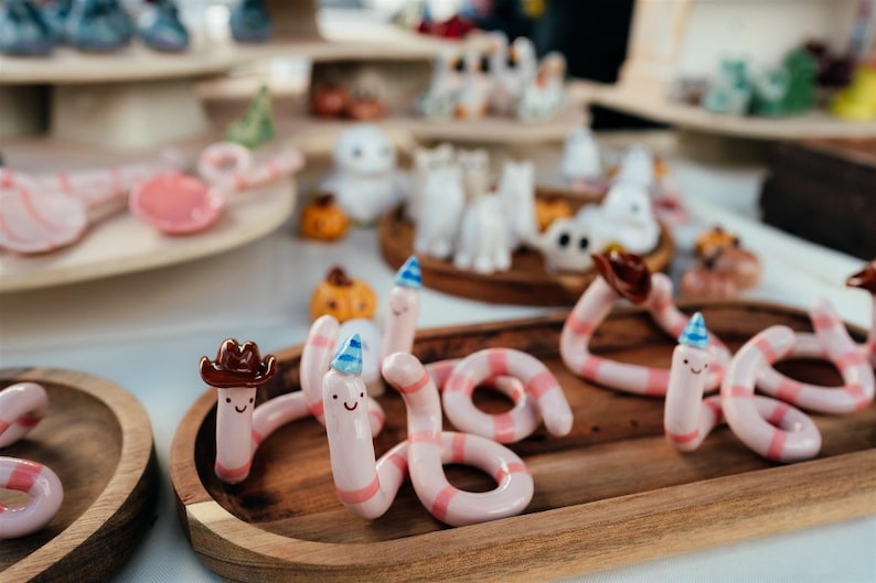 May include: A collection of small, hand-sculpted ceramic worms in light pink with darker pink stripes. Each worm features a simple smiling face and wears a tiny accessory, such as a brown cowboy hat or a blue and white patterned party hat.