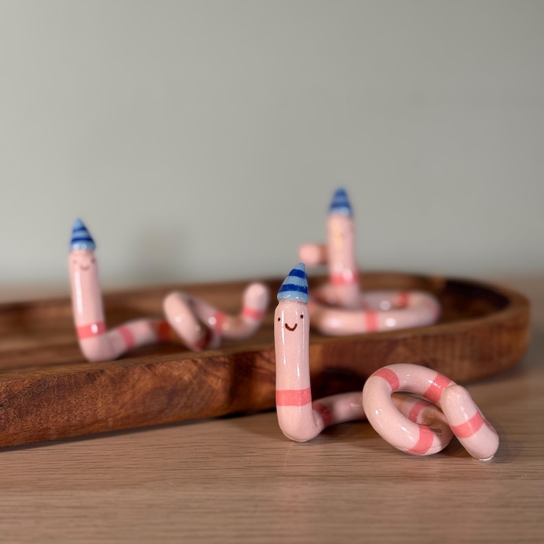May include: Three light pink ceramic worm figurines with horizontal coral-colored stripes. Each worm has a small, hand-painted smiling face and wears a tiny, pointed blue and white striped party hat on its head, with curled, flexible bodies.