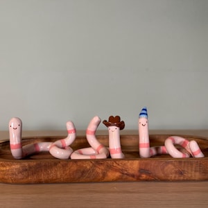 May include: Three pink and white striped ceramic worm figurines with smiling faces. One worm wears a brown cowboy hat, and another wears a blue and white striped party hat. The three figurines are arranged inside an oval wooden tray.