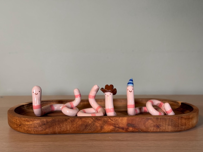 May include: Three pink and white striped ceramic worm figurines with smiling faces. One worm wears a brown cowboy hat, and another wears a blue and white striped party hat. The three figurines are arranged inside an oval wooden tray.