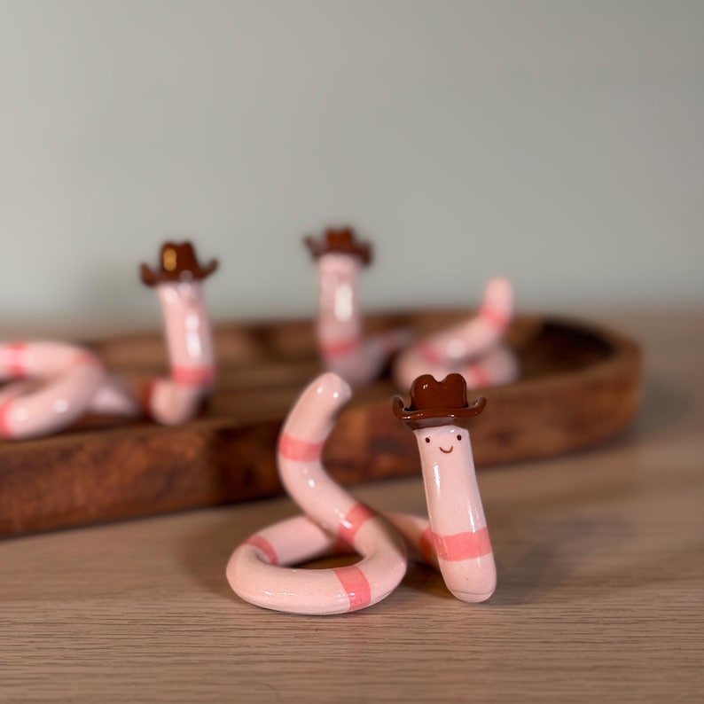 May include: Several small ceramic worm figurines, each wearing a tiny brown cowboy hat. The worms are light pink with darker pink horizontal stripes and have simple painted faces with two black dots for eyes and a small curved line for a mouth.