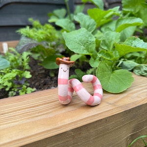 May include: A small, coiled ceramic worm figurine featuring alternating light pink and white horizontal stripes. The worm has a simple painted face with two black dots for eyes and a curved line for a mouth, topped with a brown miniature cowboy hat.