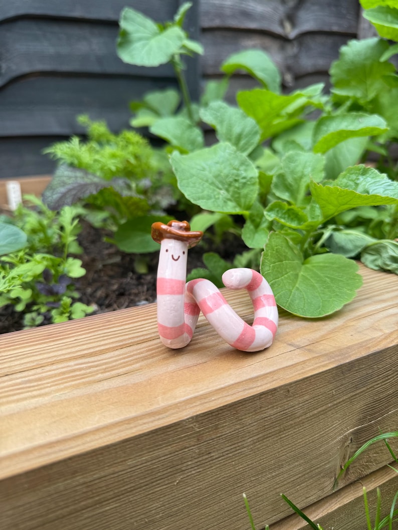 May include: A small, coiled ceramic worm figurine featuring alternating light pink and white horizontal stripes. The worm has a simple painted face with two black dots for eyes and a curved line for a mouth, topped with a brown miniature cowboy hat.