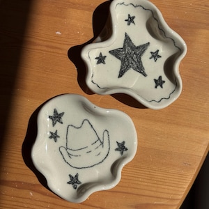 May include: Two off-white ceramic trinket dishes with irregular, wavy edges. One dish features a large central star surrounded by smaller stars, while the other displays a cowboy hat illustration surrounded by four small stars, all in black.