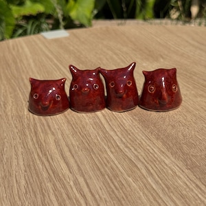 May include: Four small, glossy, dark red ceramic figurines shaped like stylised animal heads with pointed ears and simple circular eyes. Each piece has a smooth, reflective glaze and a rounded base, arranged in a horizontal row on a surface.