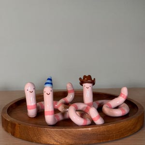 May include: A group of charming ceramic worm figurines, each with a smiling face and pink and white striped bodies. One worm wears a cowboy hat, another a party hat. The worms are displayed on a round wooden tray.