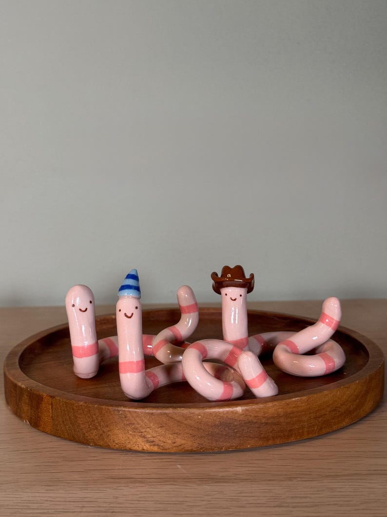May include: A set of three pink and light pink striped ceramic worms with simple painted faces. One worm wears a blue and white striped party hat, while another wears a brown cowboy hat. The worms are positioned in a curved, winding arrangement.