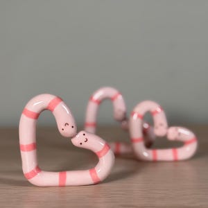 May include: Three heart-shaped sculptures in a light pink hue with horizontal stripes of a darker pink. Each heart has a smiling face. The sculptures are on a wooden surface with a grey background.