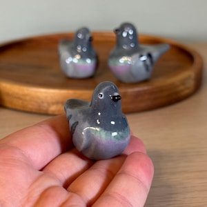 以下が含まれることがあります： 3つのセラミックハトの置物。鳥は主にグレーで、紫と緑の虹色のアクセントがあります。1つの置物は手で持たれ、他の2つは木製のトレイにあります。置物には黒いクチバシと小さくて丸い目があります。