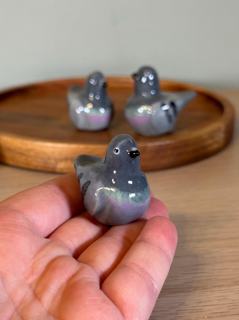 以下が含まれることがあります： 3つのセラミックハトの置物。鳥は主にグレーで、紫と緑の虹色のアクセントがあります。1つの置物は手で持たれ、他の2つは木製のトレイにあります。置物には黒いクチバシと小さくて丸い目があります。