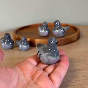 以下が含まれることがあります： 小さなセラミック製のハトの置物のコレクション。鳥は主に灰色で、羽と体に虹色のアクセントがあります。置物は木製のトレイに展示され、手に持たれています。