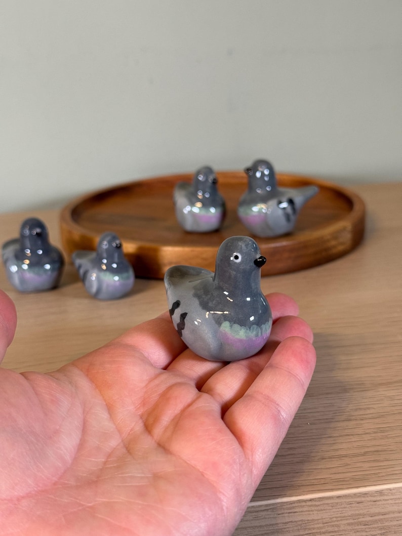 以下が含まれることがあります： 小さなセラミック製のハトの置物のコレクション。鳥は主に灰色で、羽と体に虹色のアクセントがあります。置物は木製のトレイに展示され、手に持たれています。