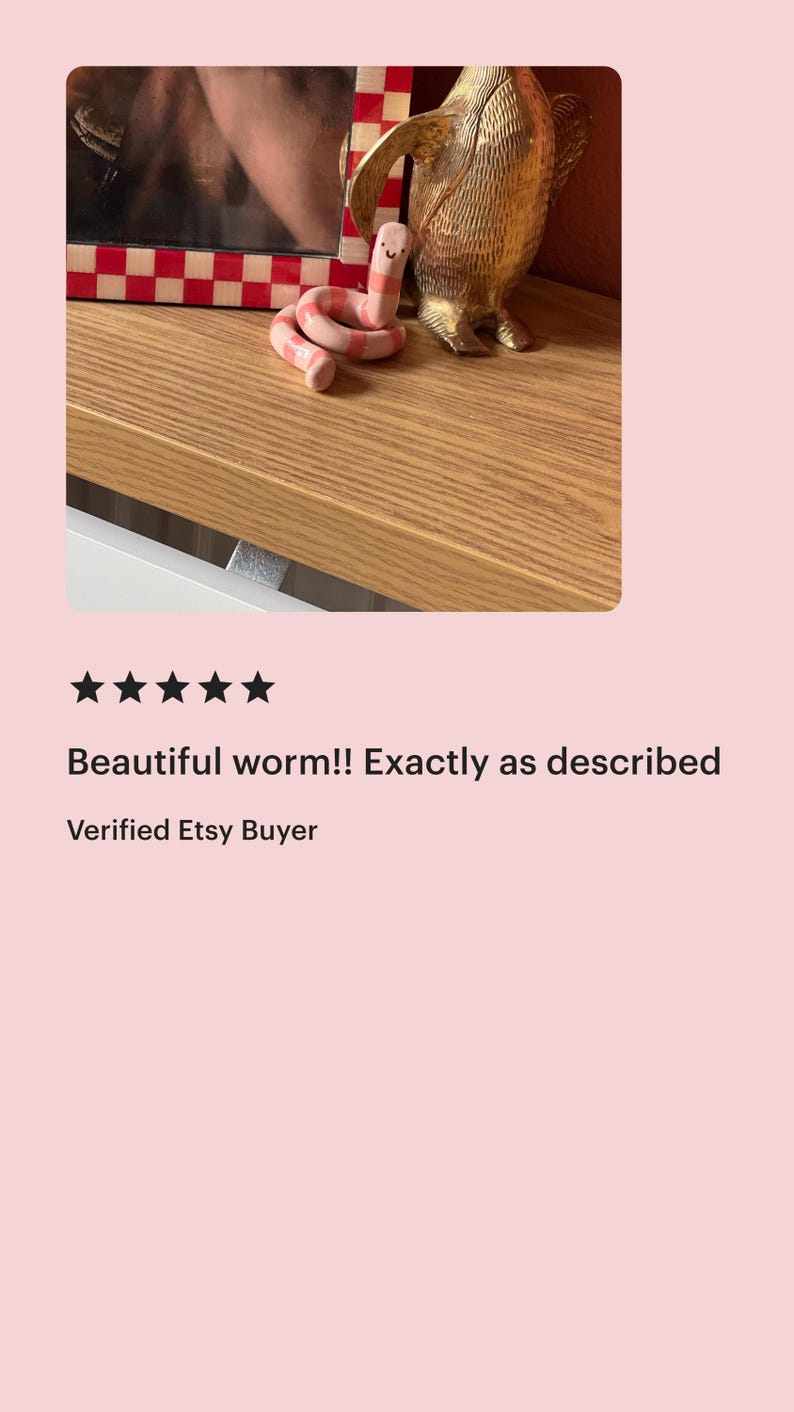 May include: A small, coiled ceramic worm figurine featuring alternating light pink and white horizontal stripes, with a simple hand-painted face consisting of two black dots for eyes and a small curved line for a mouth, positioned on a wooden surface.