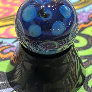 May include: A glass marble with a dark blue core and light blue spots. The marble has a glittery, cosmic design and sits on a black, glossy base. The marble's surface reflects light, creating a captivating visual effect.