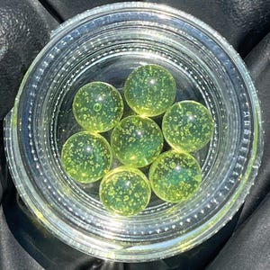 May include: A clear glass jar filled with seven translucent, yellow-green spheres. The spheres have a bubbly texture and are evenly spaced within the jar. The jar is sitting on a dark surface.