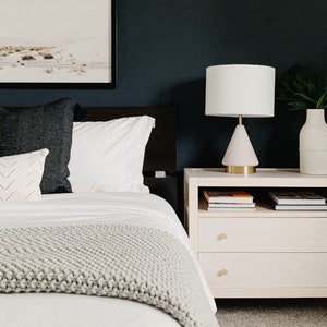 May include: A white nightstand with two drawers and a shelf, featuring a white lamp with a white shade, a white vase with green leaves, and two books on the shelf. The nightstand is next to a bed with white bedding and a gray knitted throw blanket.