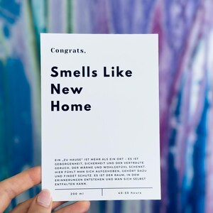 May include: A white card with the text "Congrats, Smells Like New Home" in black bold font. The card also includes the text "EIN ZU HAUSE' IST MEHR ALS EIN ORT - ES IST GEBORGENHEIT, SICHERHEIT UND DER VERTRAUTE GERUCH, DER W&Auml;RME UND WOHLGEF&Uuml;HL SCHENKT. HIER F&Uuml;HLT MAN SICH AUFGEHOBEN, GEH&Ouml;RT DAZU UND FINDET SCHUTZ. ES IST DER RAUM, IN DEM ERINNERUNGEN ENTSTEHEN UND MAN SICH SELBST ENTFALTEN KANN." in smaller black font. The card also includes the text "200 ml" and "40-50 hours" in black font.