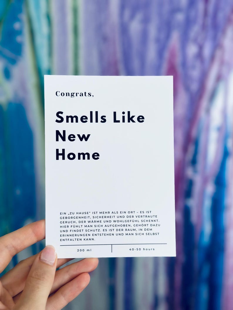 May include: A white card with the text "Congrats, Smells Like New Home" in black bold font. The card also includes the text "EIN ZU HAUSE' IST MEHR ALS EIN ORT - ES IST GEBORGENHEIT, SICHERHEIT UND DER VERTRAUTE GERUCH, DER W&Auml;RME UND WOHLGEF&Uuml;HL SCHENKT. HIER F&Uuml;HLT MAN SICH AUFGEHOBEN, GEH&Ouml;RT DAZU UND FINDET SCHUTZ. ES IST DER RAUM, IN DEM ERINNERUNGEN ENTSTEHEN UND MAN SICH SELBST ENTFALTEN KANN." in smaller black font. The card also includes the text "200 ml" and "40-50 hours" in black font.