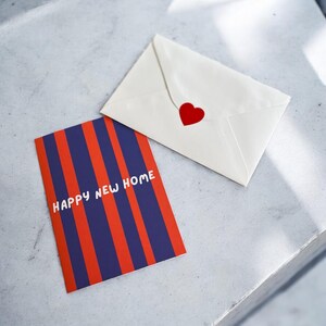 May include: A red and blue striped card with the text "Happy New Home" in white. A white envelope with a red heart sticker is next to the card.