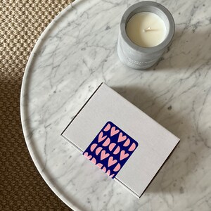 May include: A white candle in a gray concrete jar with the text "Sandalwood Myrrh" on the side. The candle is sitting on a white marble surface with a white box with a pink and blue heart pattern on top.